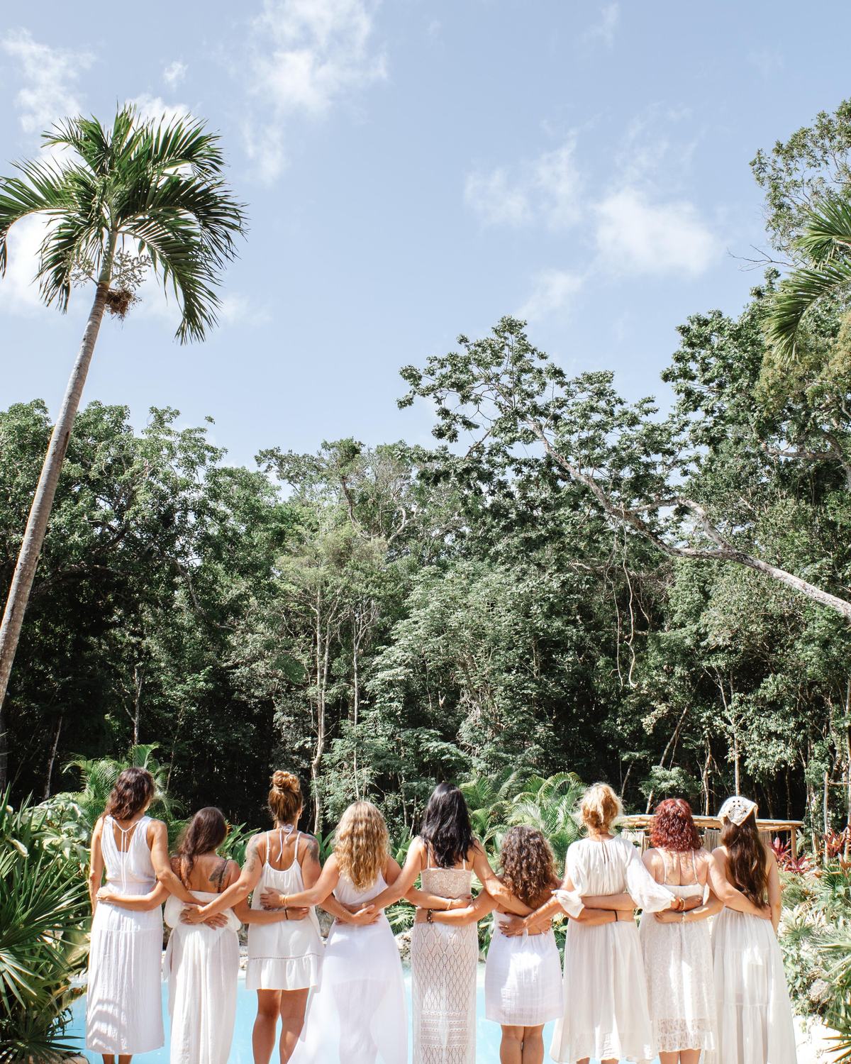 A circle of women in white standing arm in arm, facing the Tulum jungle and open sky