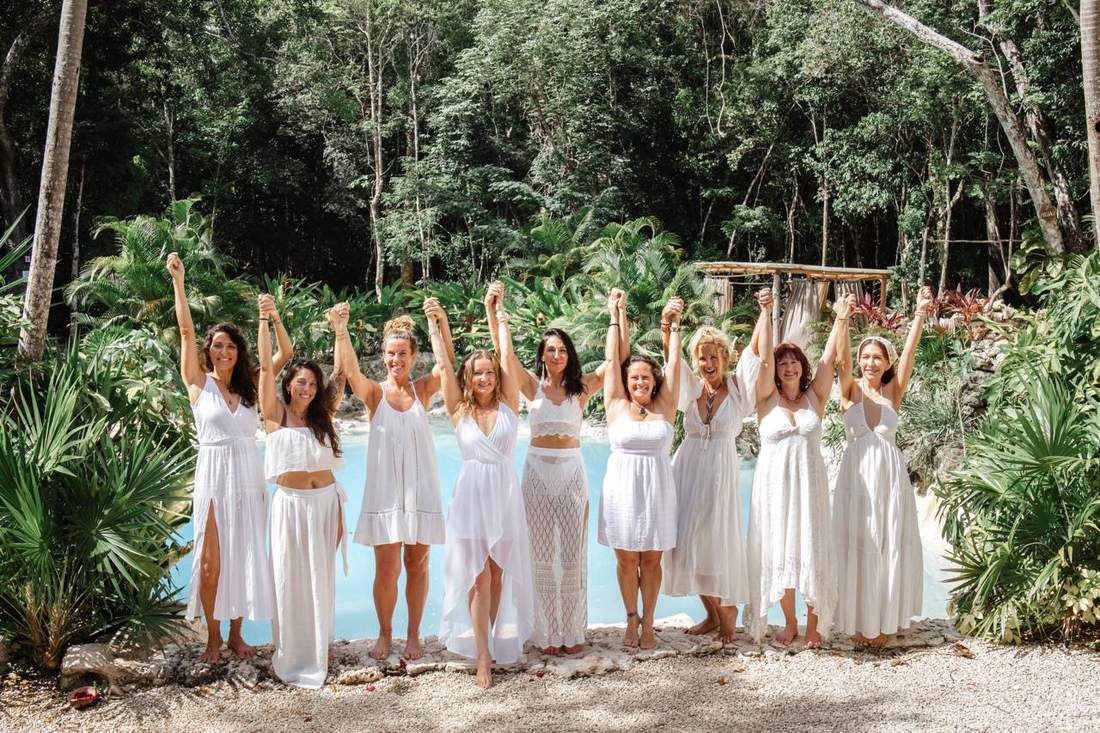 Nine women in white dresses holding raised hands before a turquoise cenote in the Tulum jungle