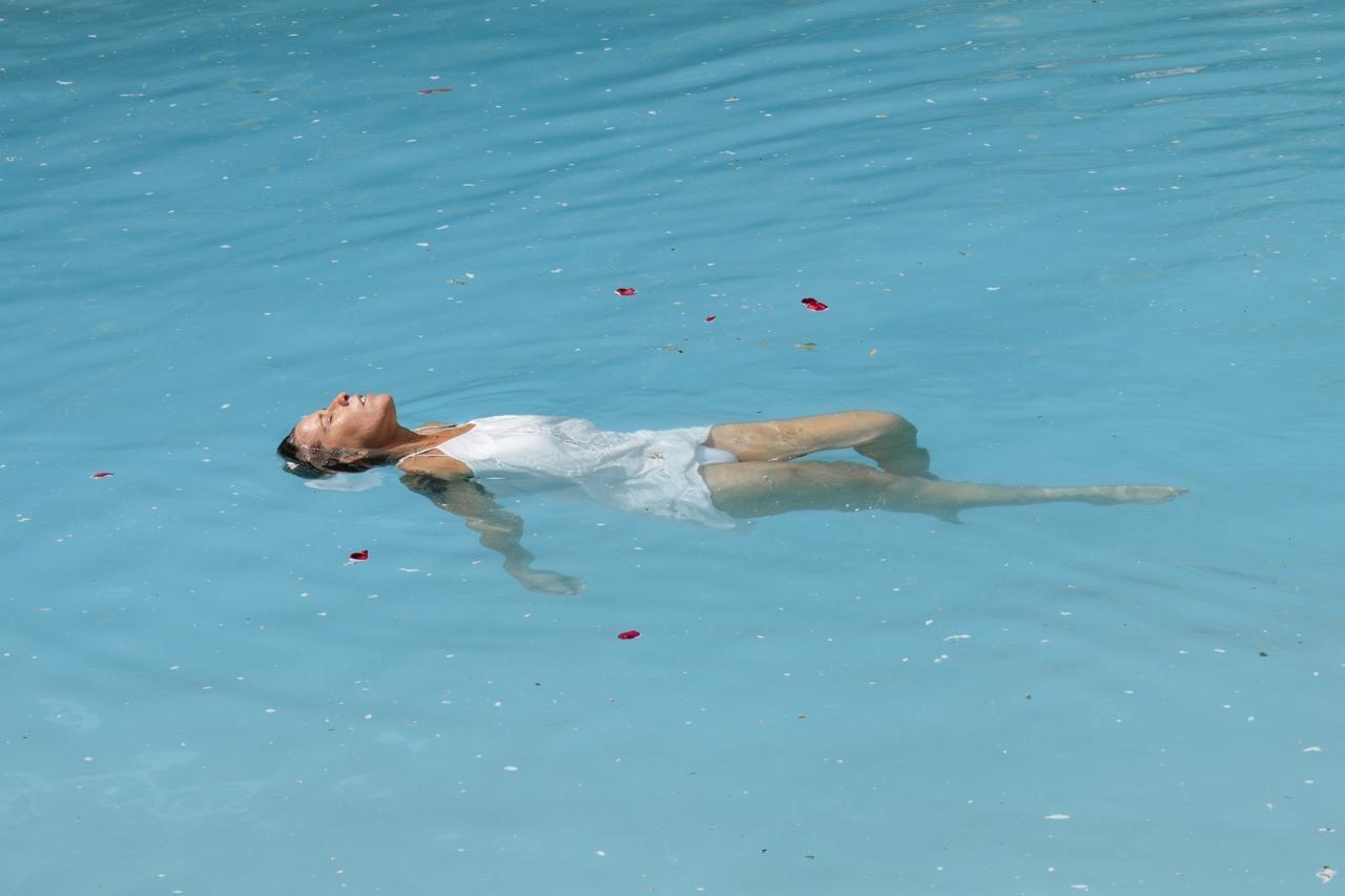 A woman in white floating in stillness on turquoise cenote water, rose petals drifting around her