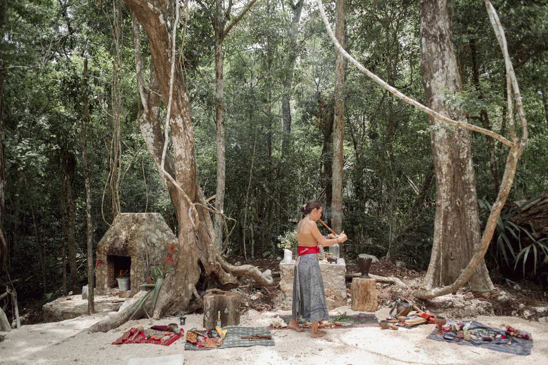 A ceremonial guide playing a wooden flute beside the sacred fire and stone altar, jungle trees rising all around