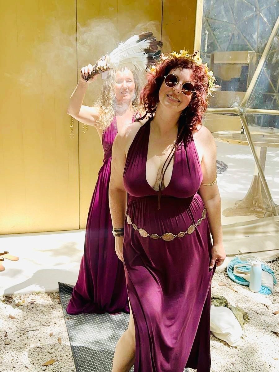 Two women in plum priestess gowns, one fanning copal smoke with a feather fan before the gold dome