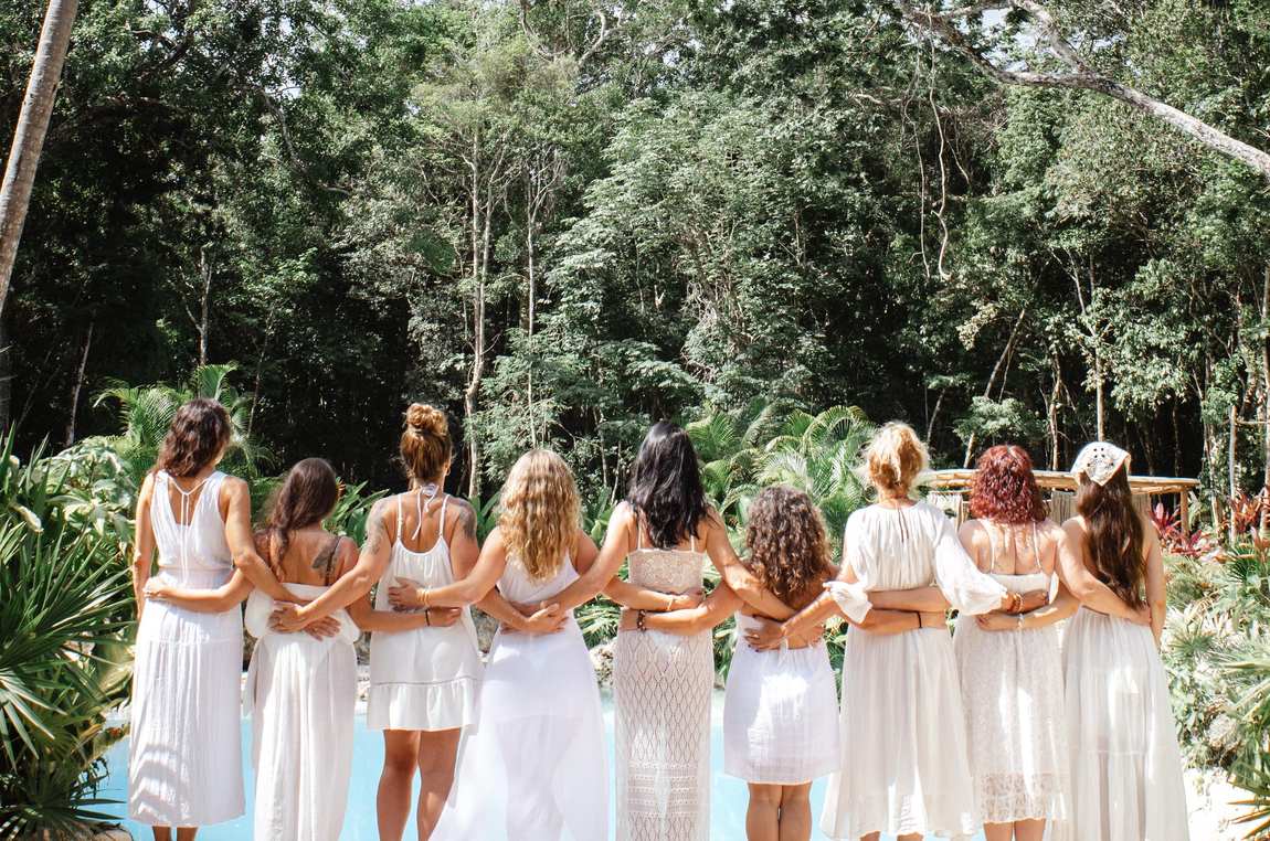 The circle of sisters in white standing arm in arm before the turquoise cenote and jungle